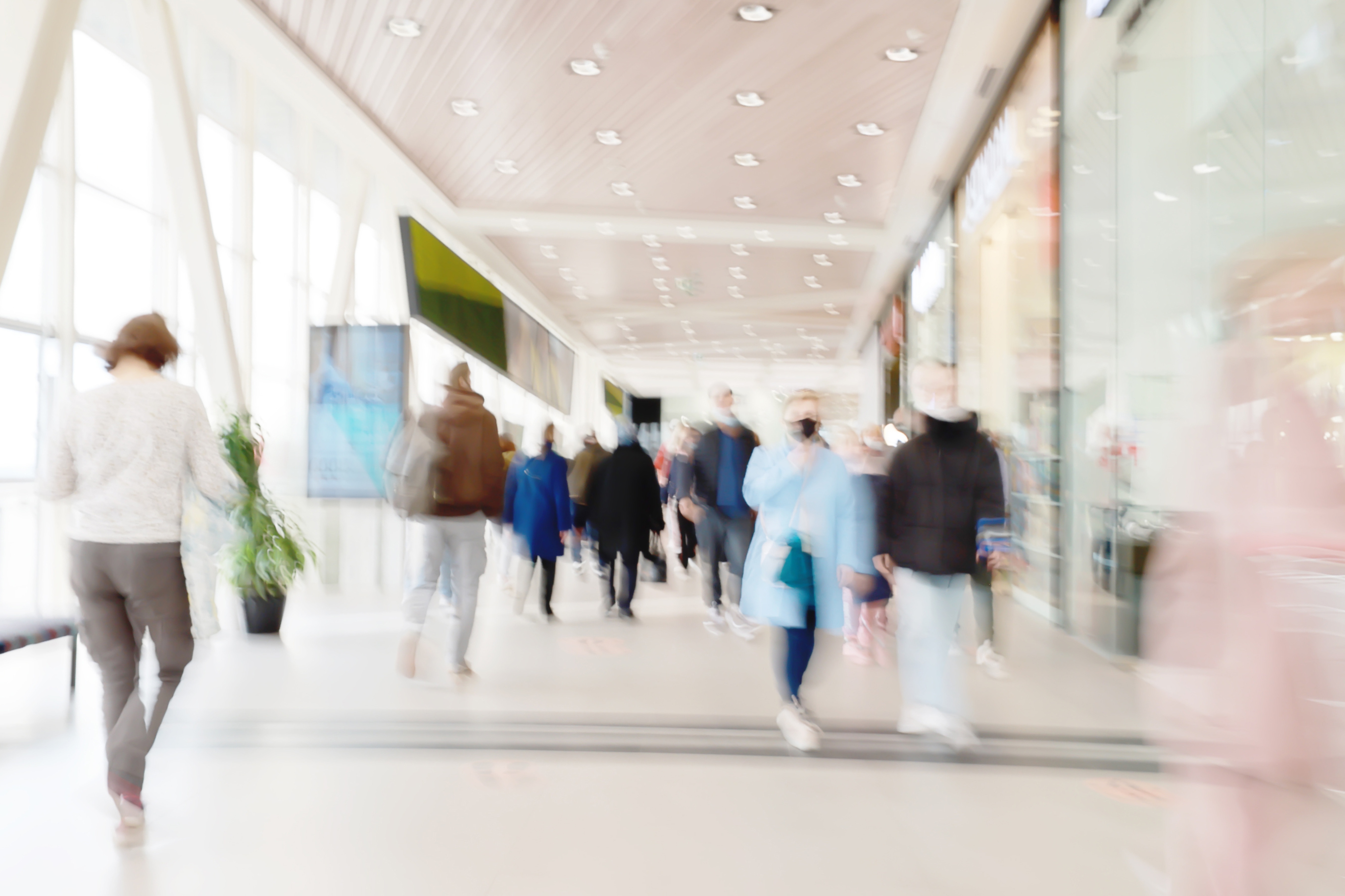 People walking through a bright indoor hallway with glass walls and shops.