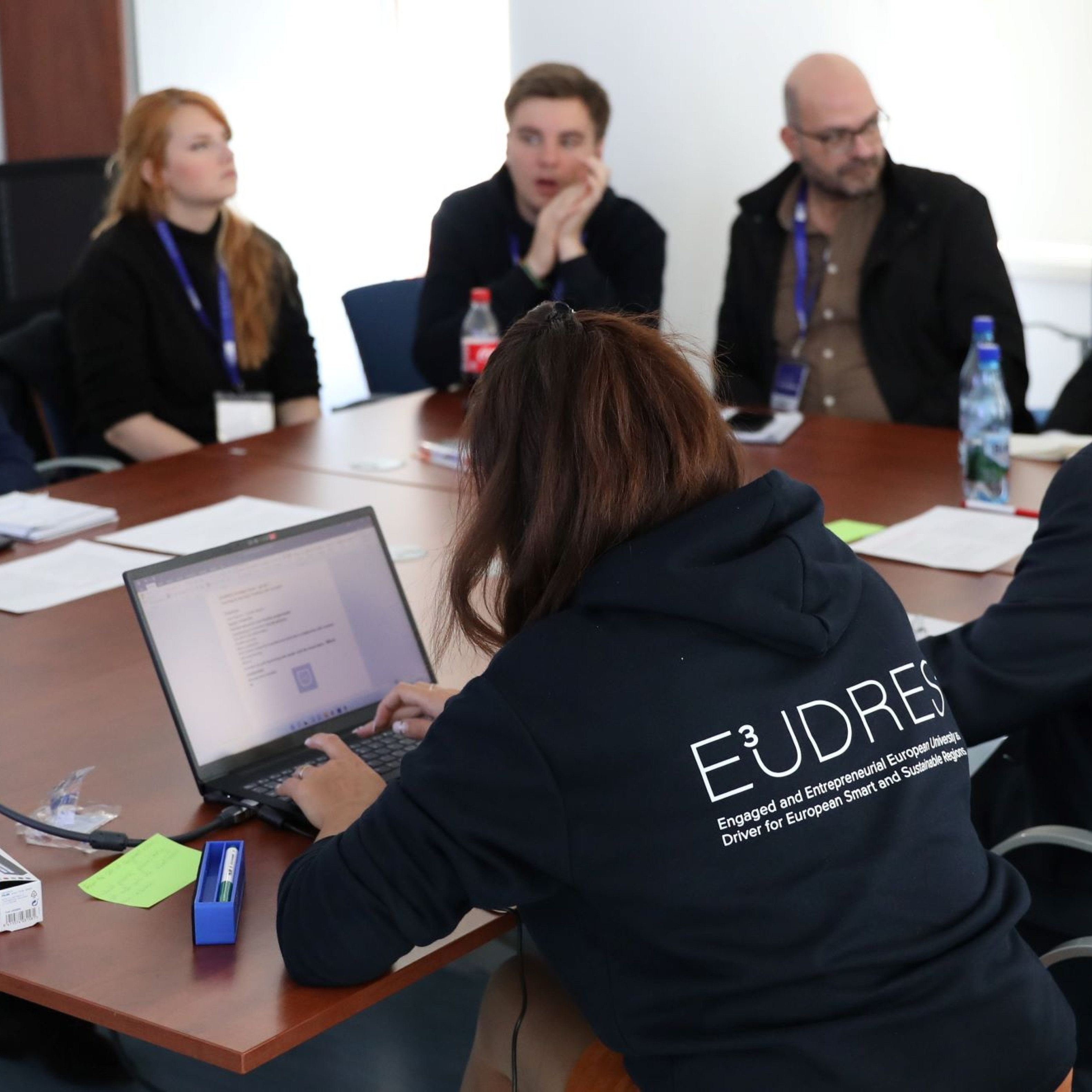 People sitting around a conference table during a meeting, one wearing an E³UDRES² hoodie.
